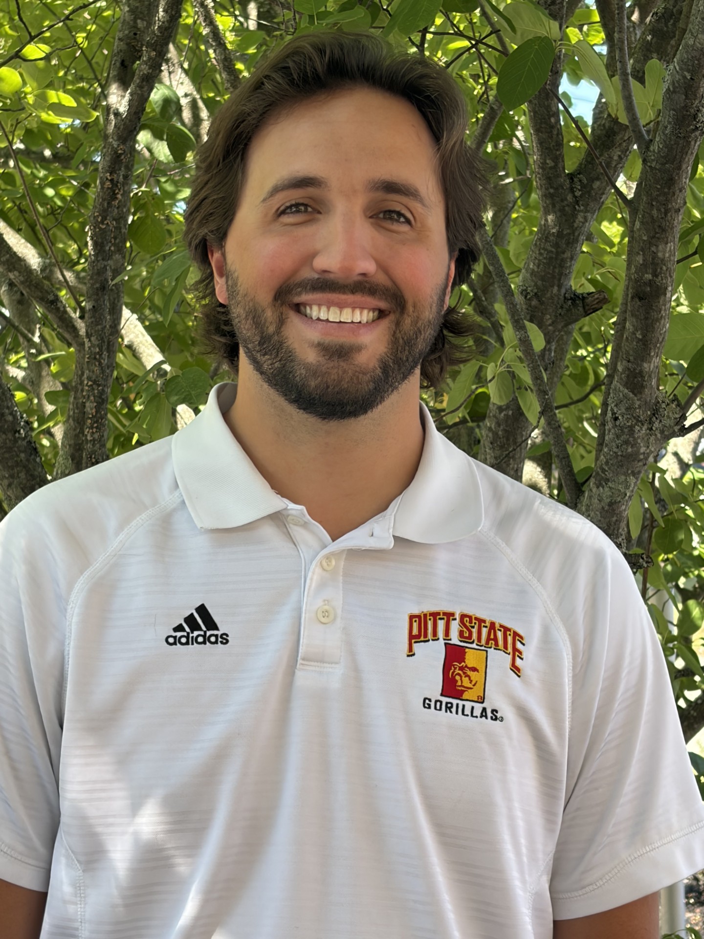 Man smiling outdoors wearing a white Pitt State Gorillas polo with Adidas logo, green leaves in the background behind tree branches.