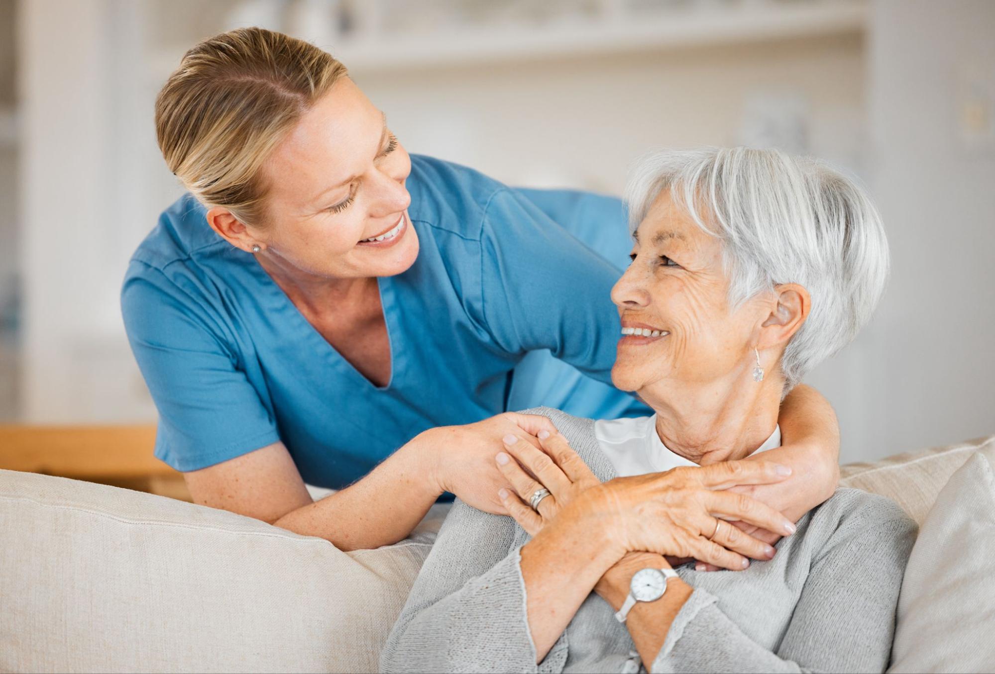 Caregiver providing companion care for elderly woman, sharing a warm hug on her couch.