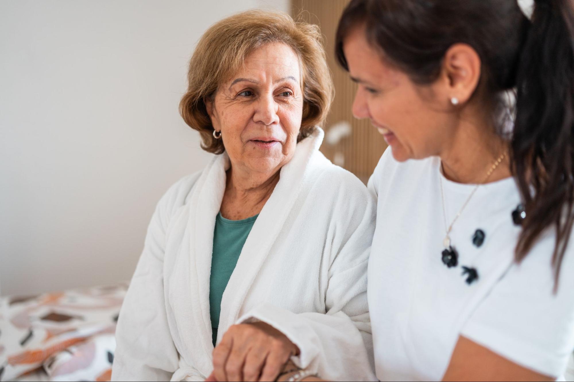 Smiling caregiver sits with an older woman in a bathrobe during bathing assistance for elderly.