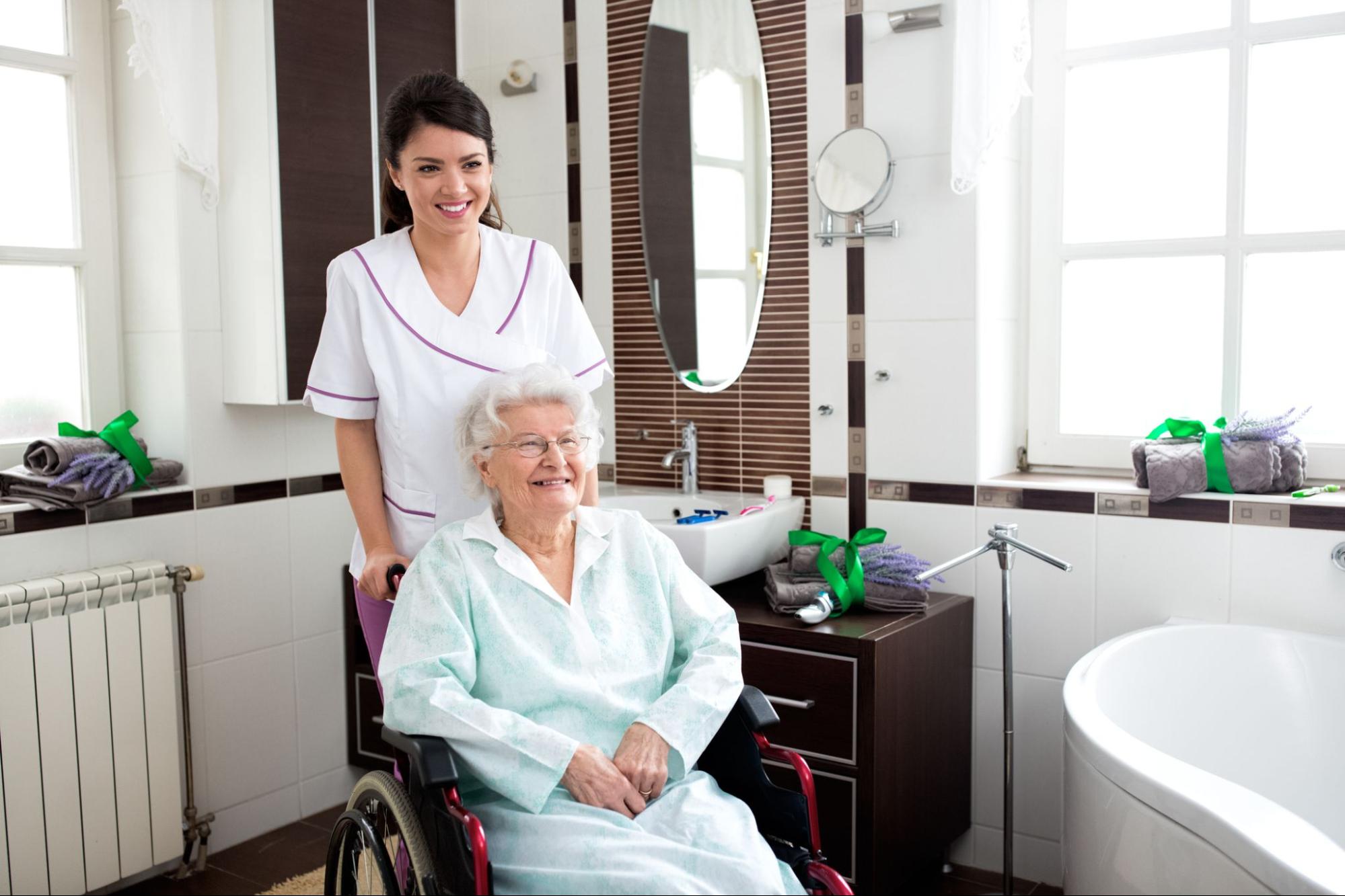 Caregiver wheels a smiling older woman into her bathroom, providing help with toileting at home.