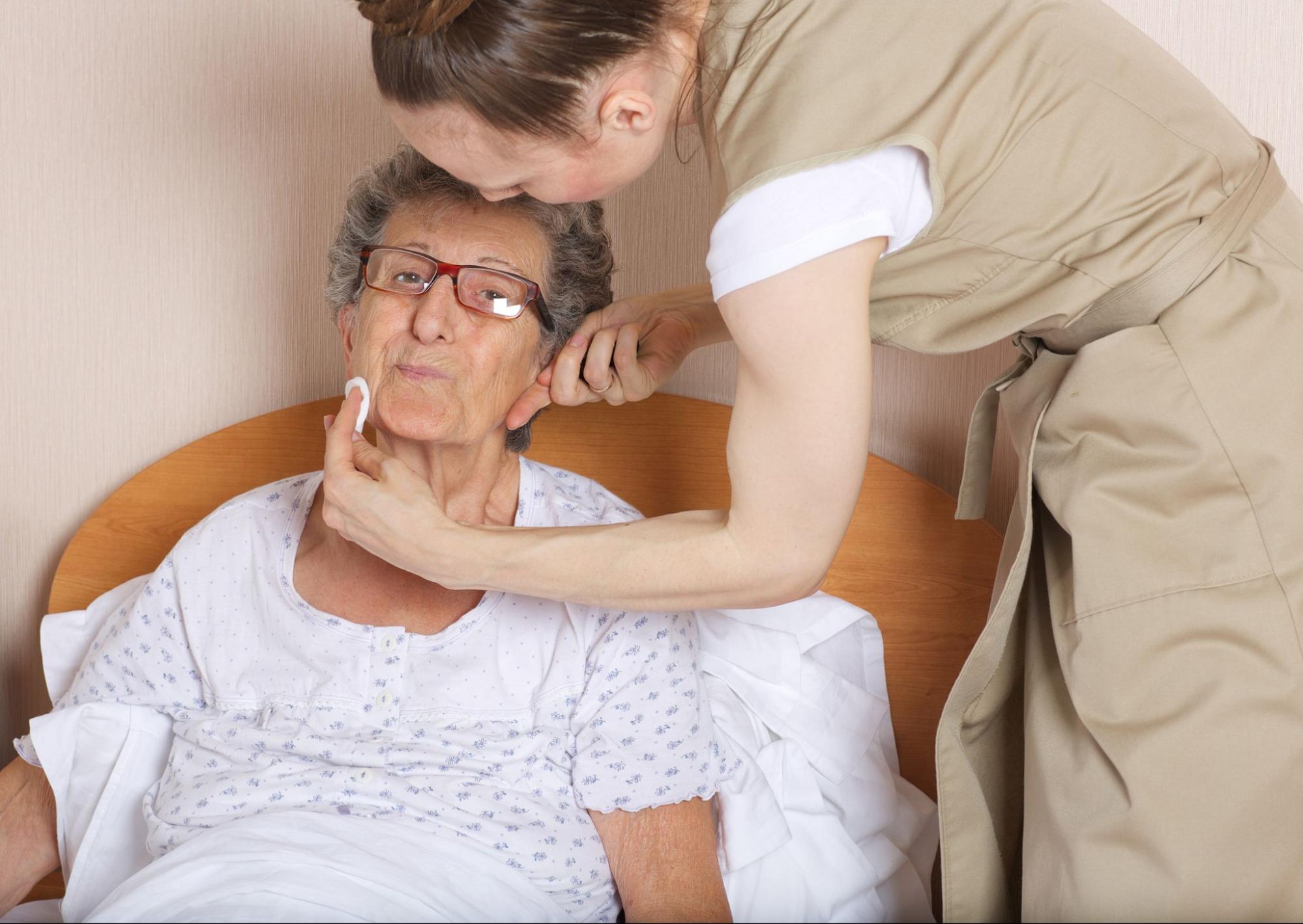 Caregiver gently cleaning an older woman’s face in bed during personal hygiene assistance.
