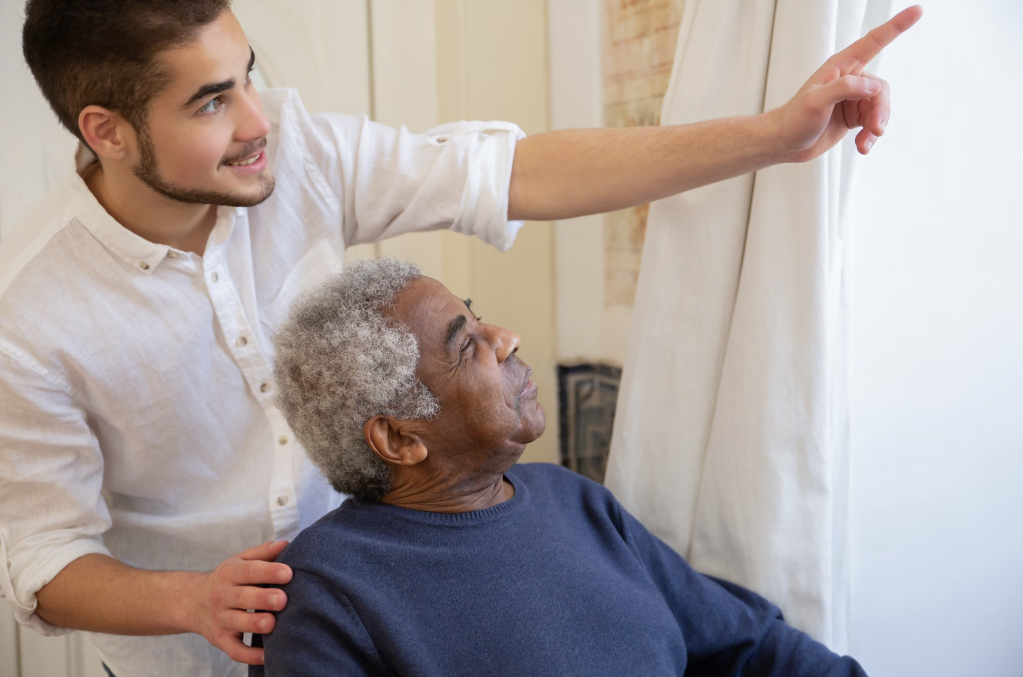 Young caregiver pointing out the window with an older man during in-home respite care.
